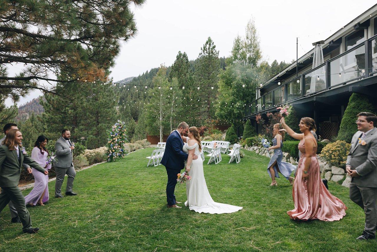 Autumn and Peter kiss on the lawn while their wedding party dances around them, with the Tannenbaum Event Center behind
