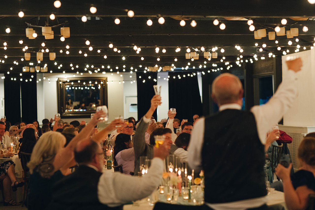A raised glass at the front of the room, and every guest raising a glass back under the string lights