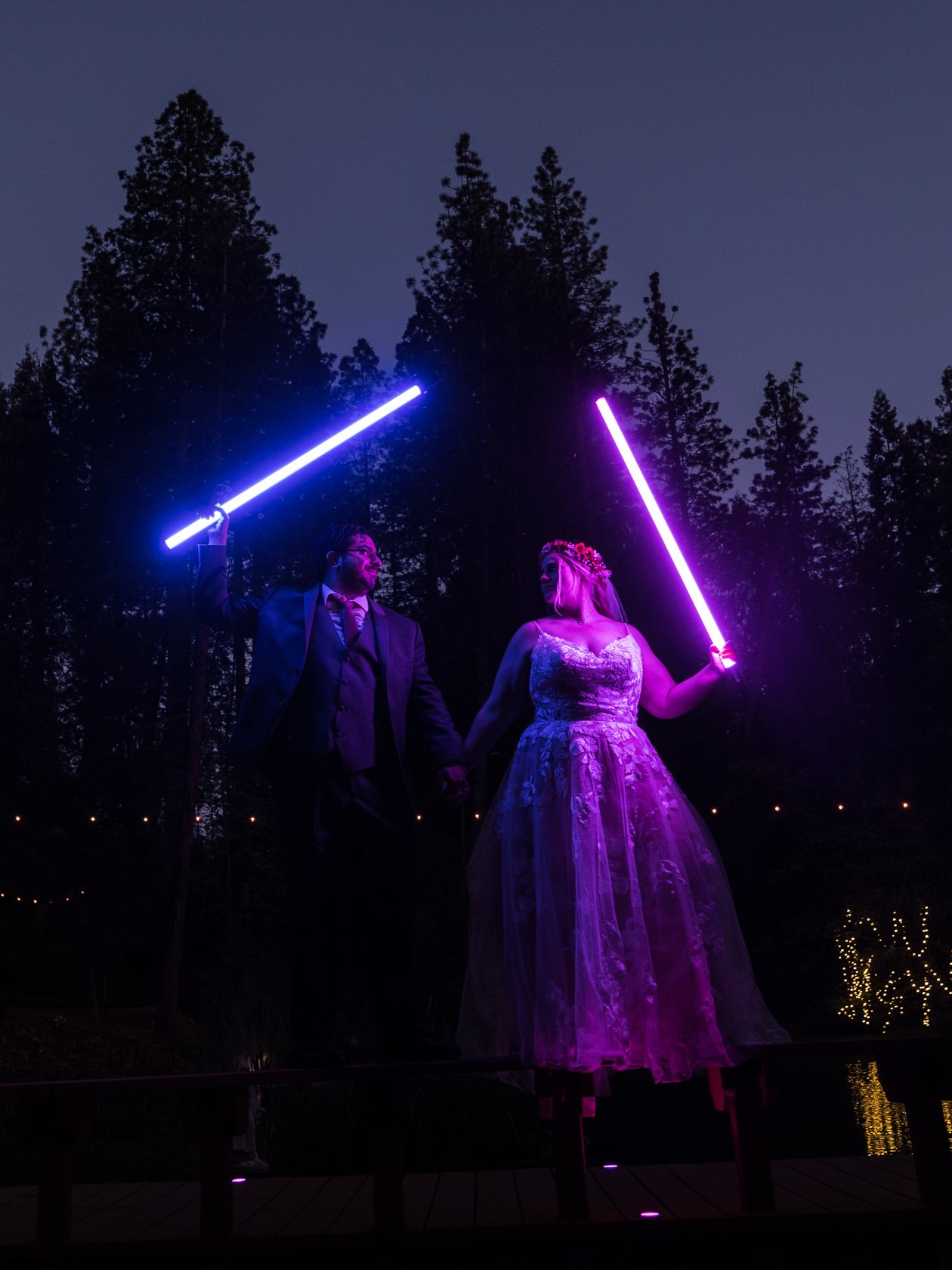 Abney and Chris raise glowing blue and purple lightsabers under the pines at dusk on their Star Wars wedding day