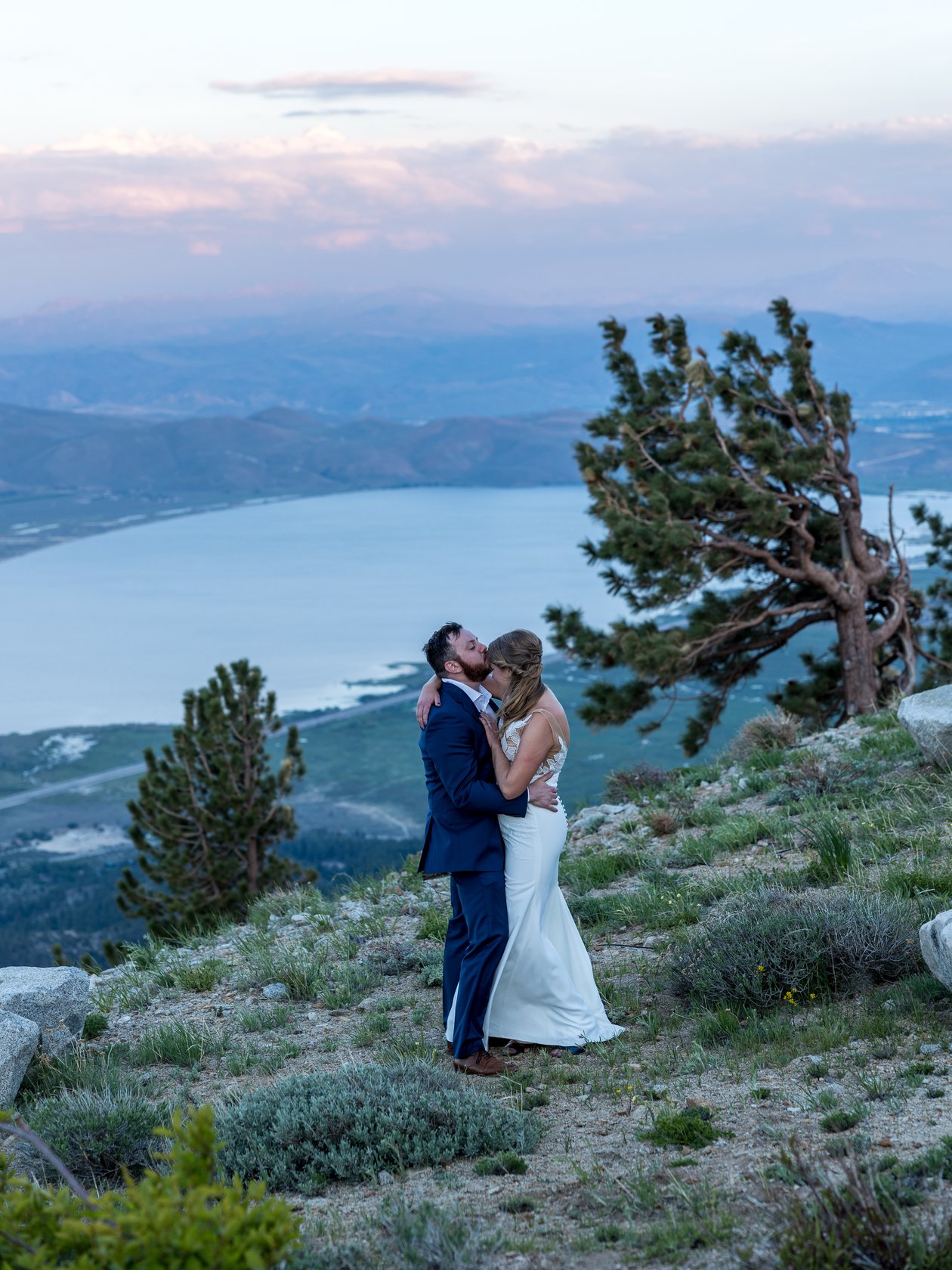 Ali and Robert kiss under pink sunset clouds beside a windswept pine above Washoe Lake