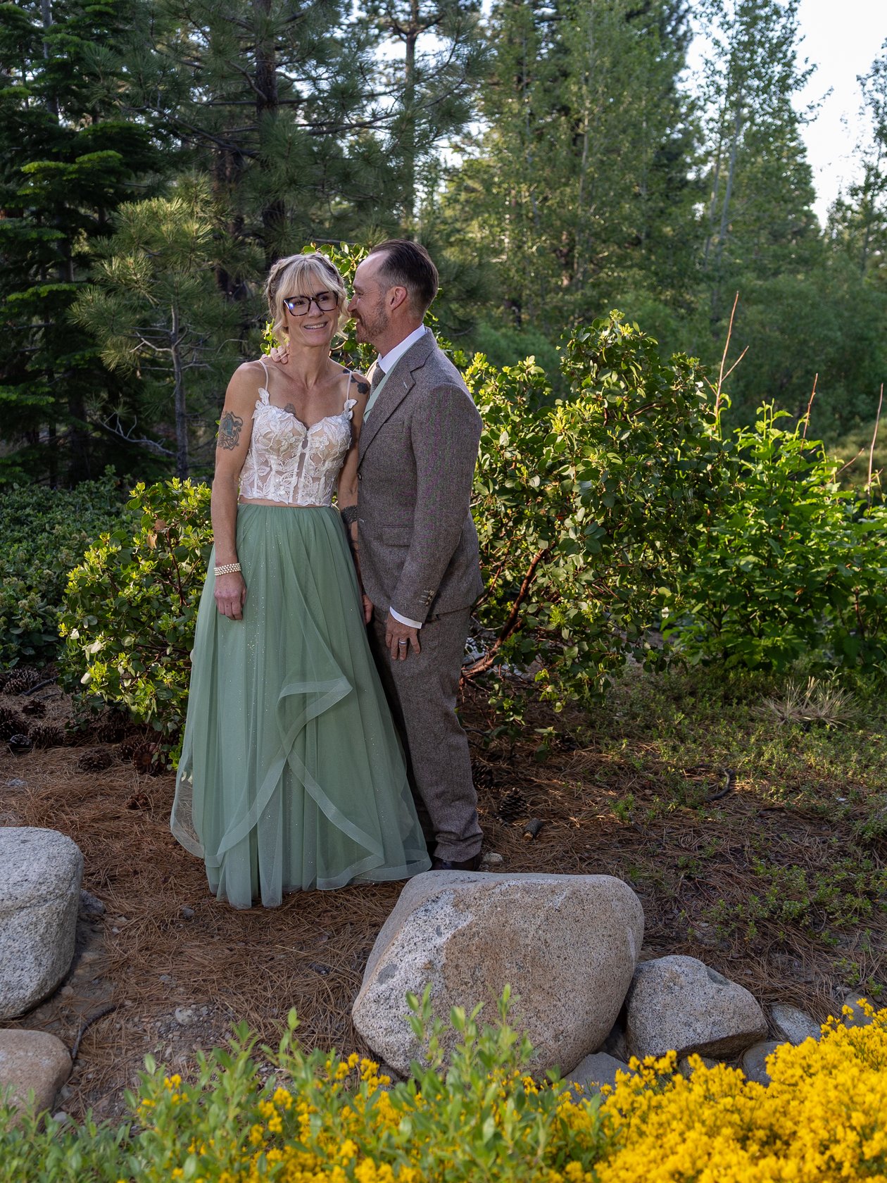 Amy and Justin stand close among the pines, yellow wildflowers and granite boulders at their feet