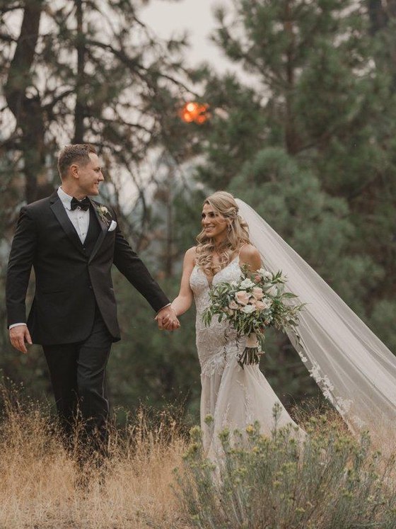 Angela and Stephen walk hand in hand through a meadow among the pines, her long veil trailing behind, photo by Composing Reality Photography