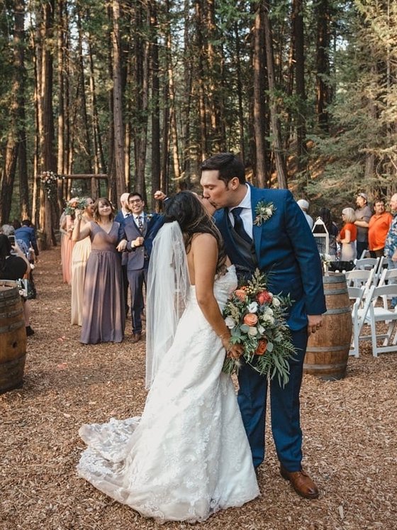 Hannah and Kyle kiss in the forest aisle between wine barrels and lanterns, photo by Composing Reality Photography