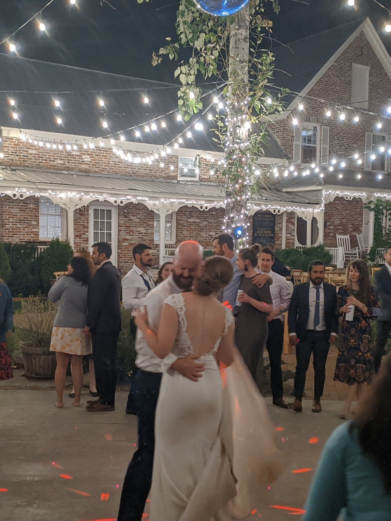 Lauren and Mark embrace during their last dance under string lights and a disco ball, the brick house glowing behind them