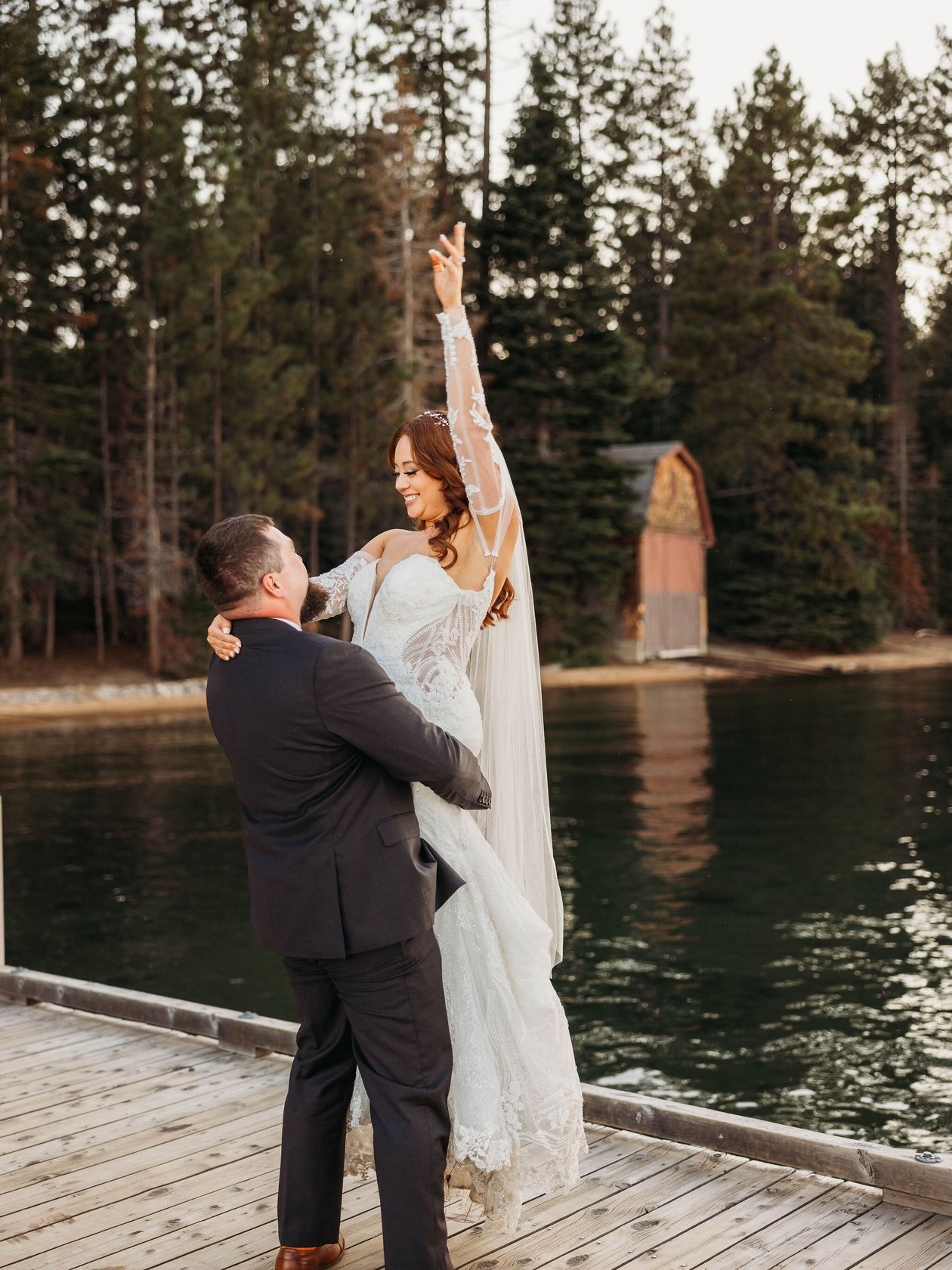 Tom lifts Remy on the dock at Valhalla as she raises one arm, the red boathouse and the lake behind them