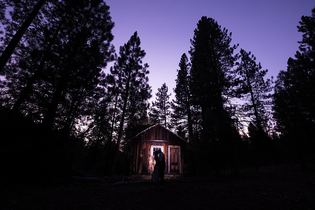 Correen and Kellen silhouetted in the lit doorway of a small cabin under tall pines at dusk