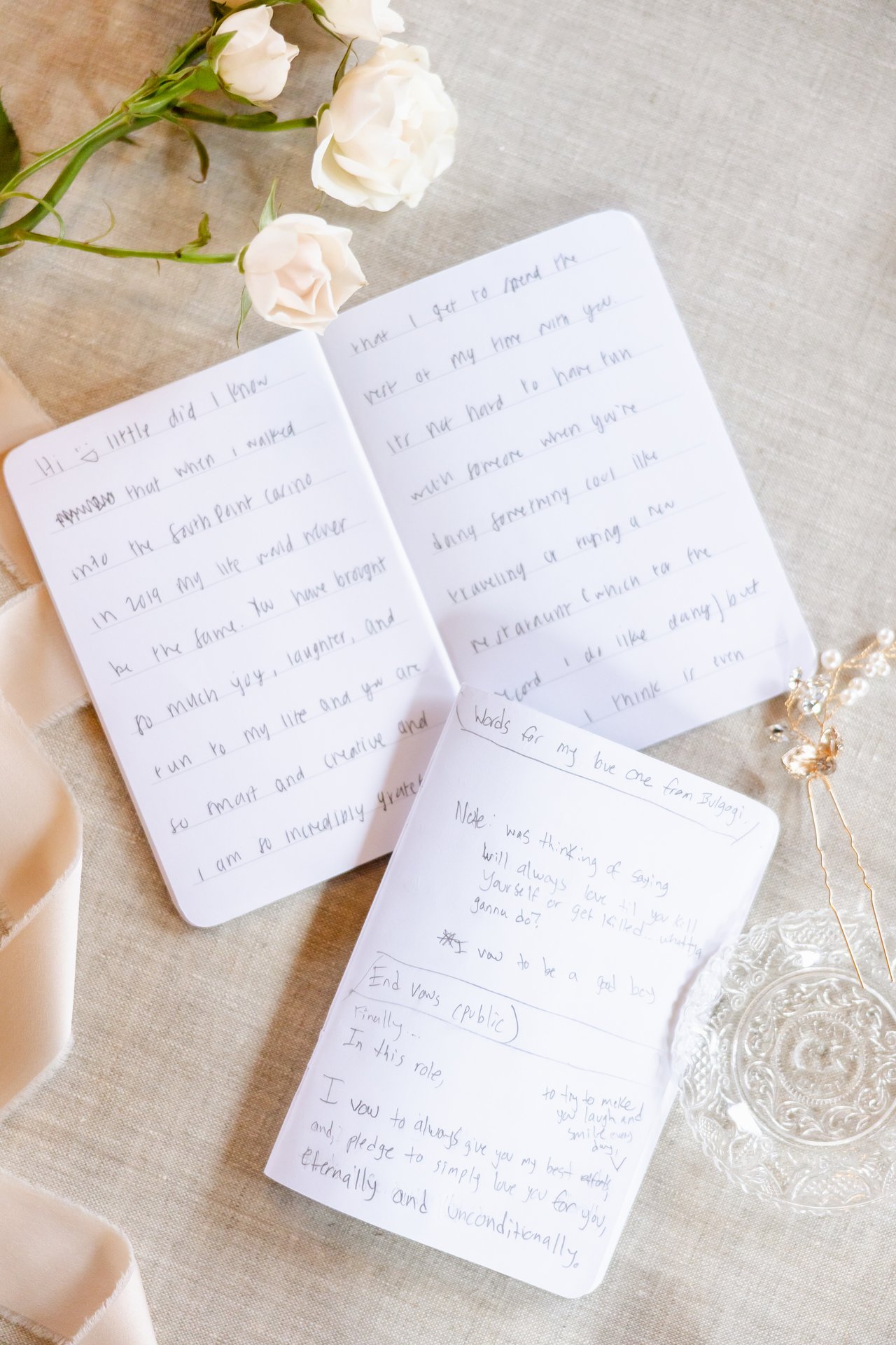 Two handwritten vow books open on linen beside white roses