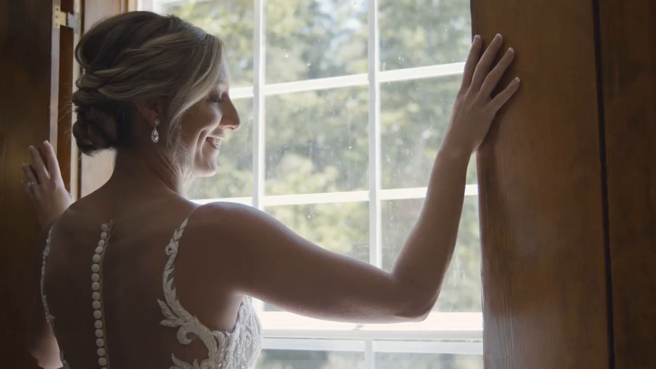 Danielle smiles out the window, the buttons and lace of her open back gown in the light.