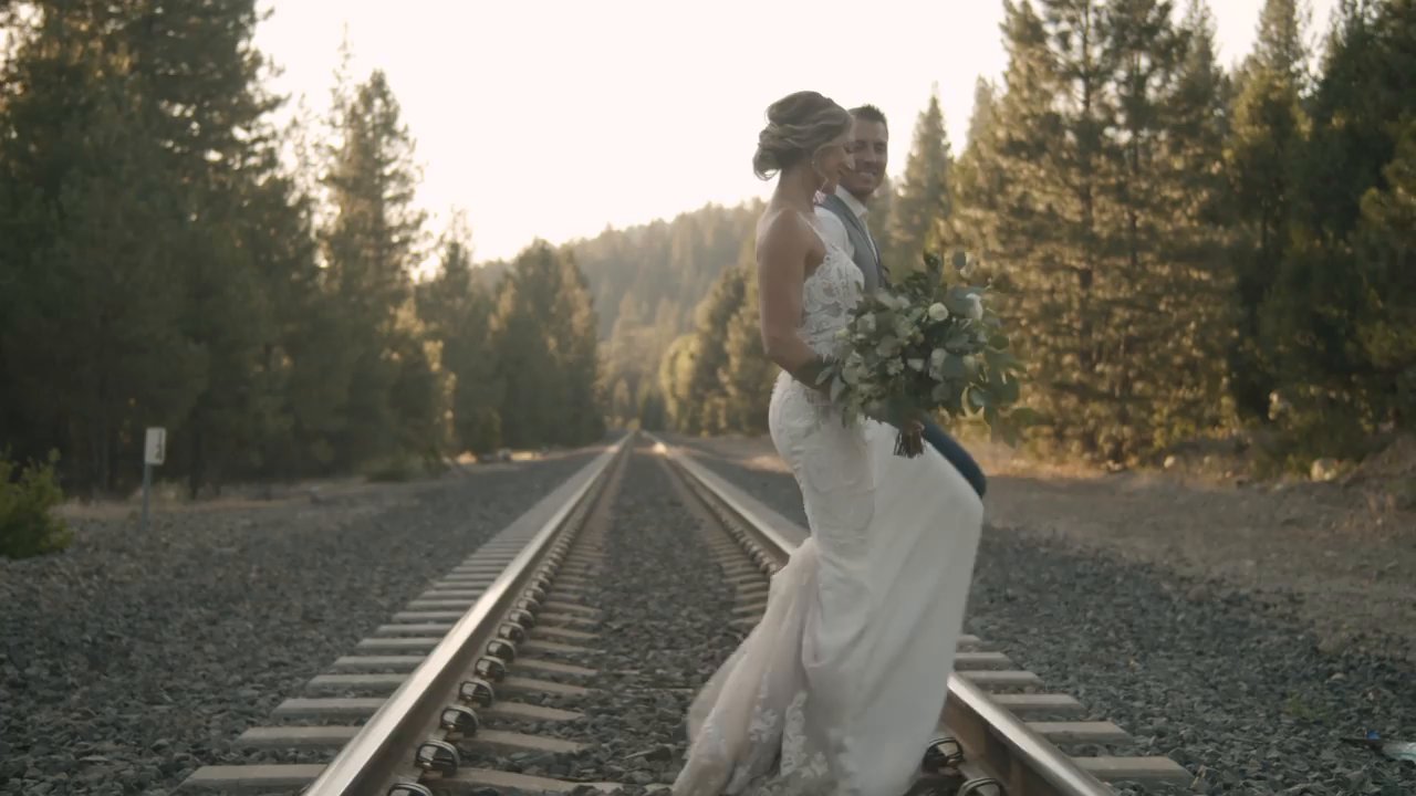 Danielle stands on the railroad tracks with her bouquet as Scott holds her.