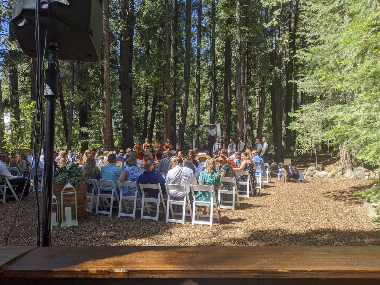 The forest ceremony from my DJ booth, guests in white chairs under the pines and the wedding party at the wooden arch