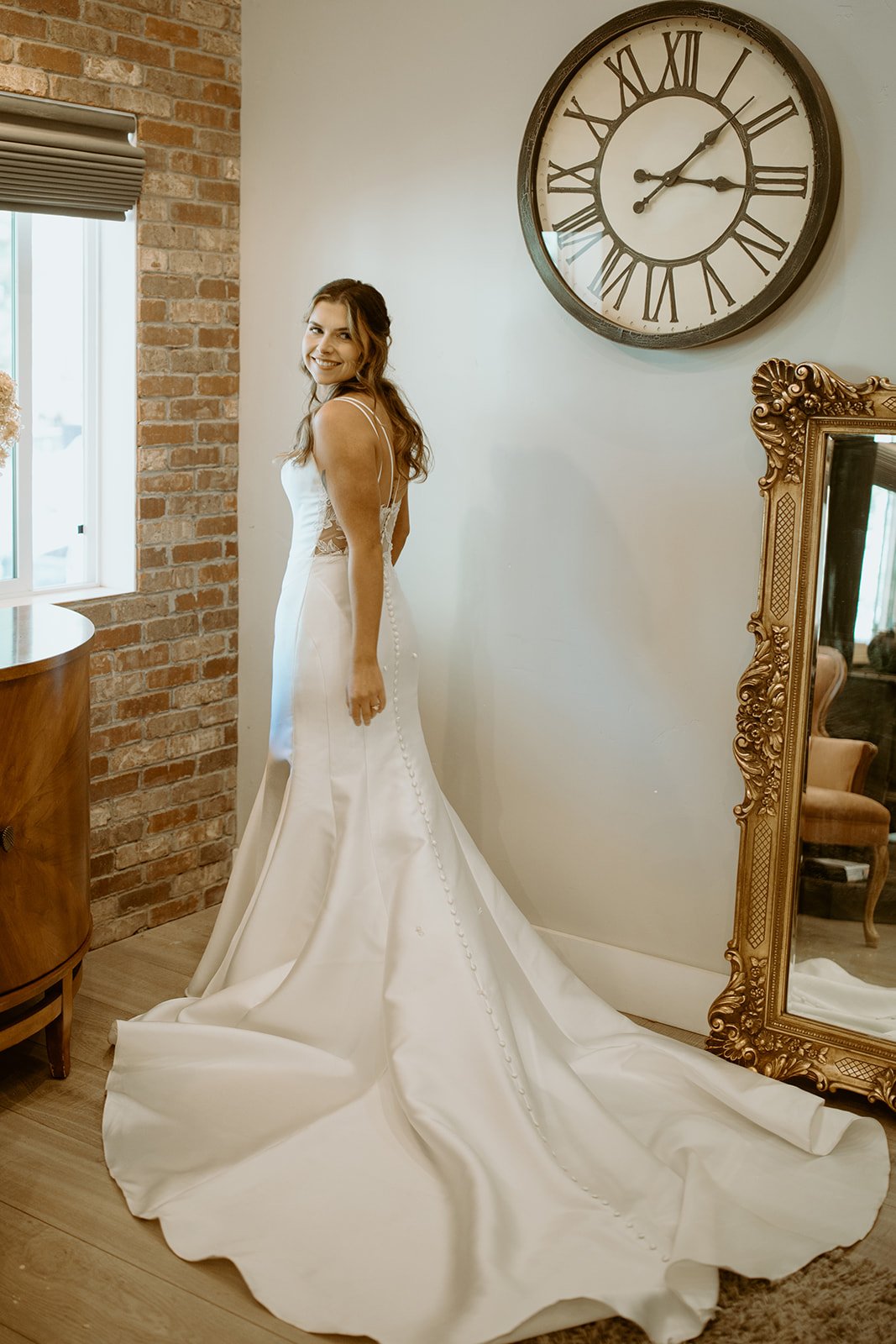 Emily looks back over her shoulder by the gold mirror, her train spread on the floor