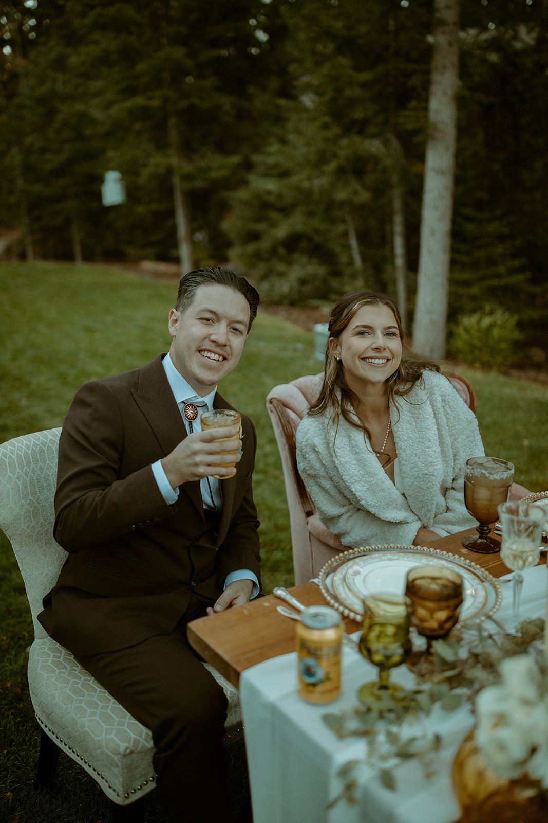 Devin raises his glass beside Emily at their table on the lawn