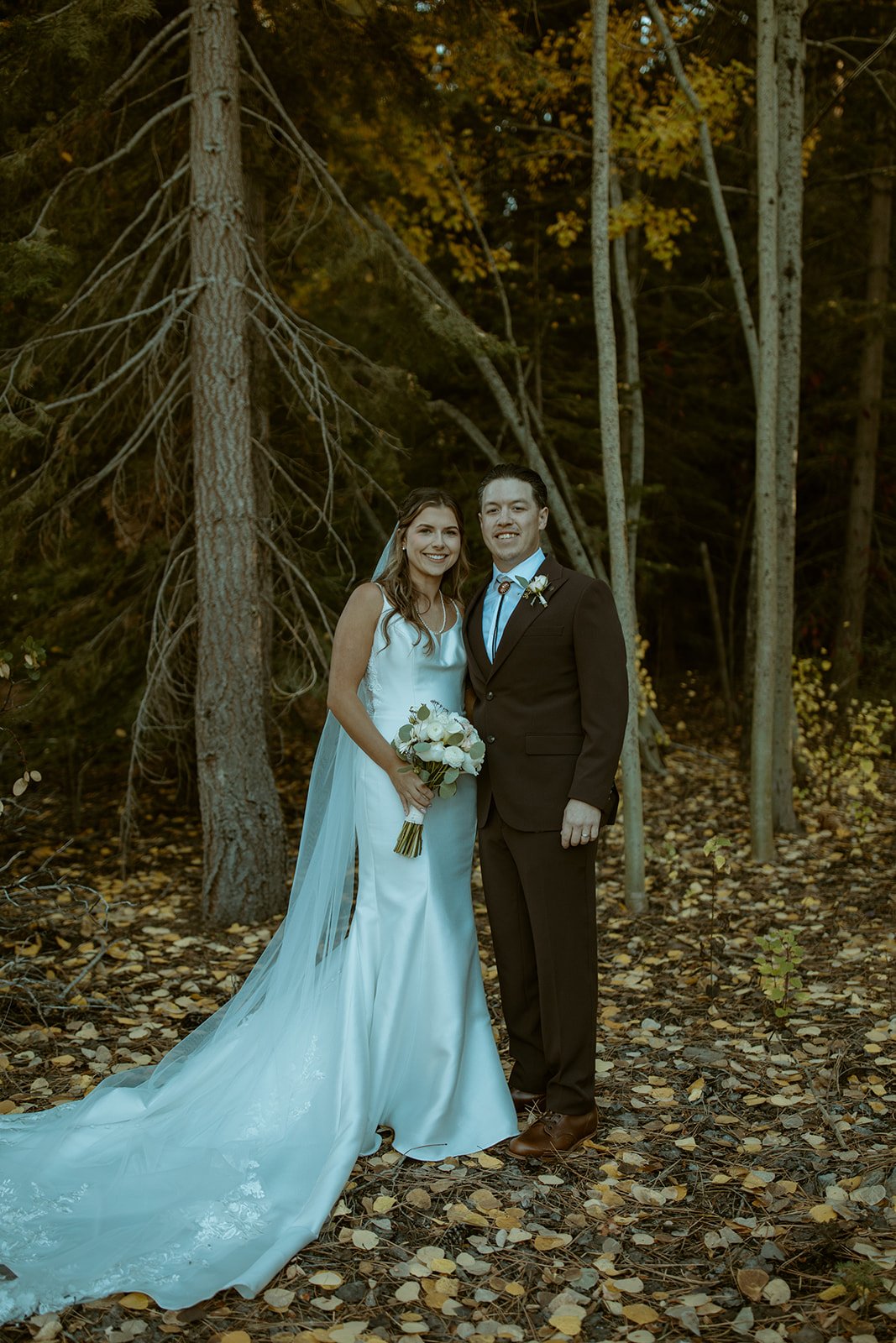 Emily and Devin smile among the trees, her train across the leaves