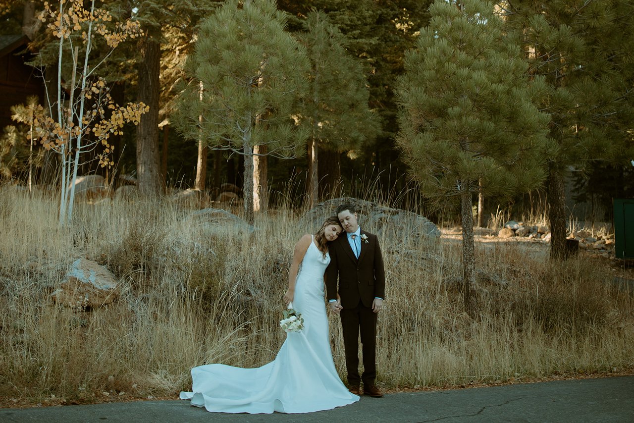 Emily rests her head on Devin's shoulder in a meadow of tall grass and pines