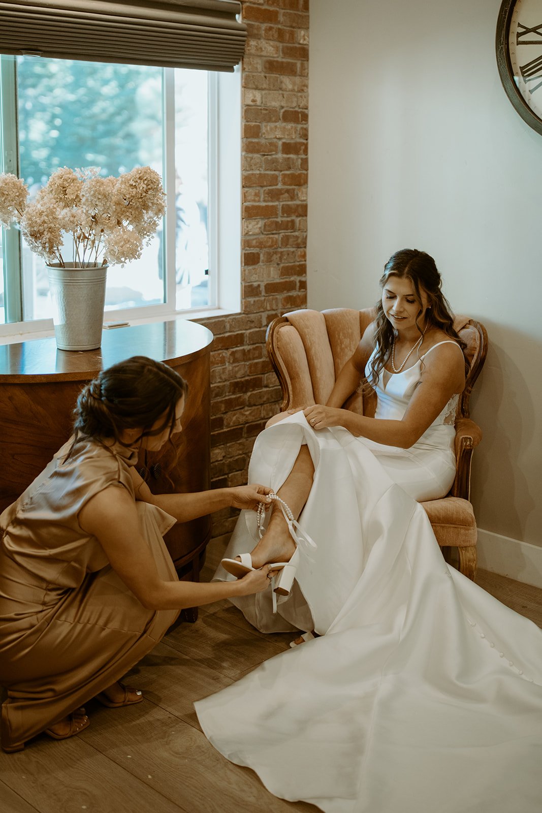 A bridesmaid helps Emily into her shoes beside the window