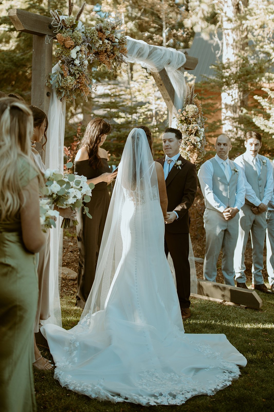 Emily's long veil at the floral arch, facing Devin