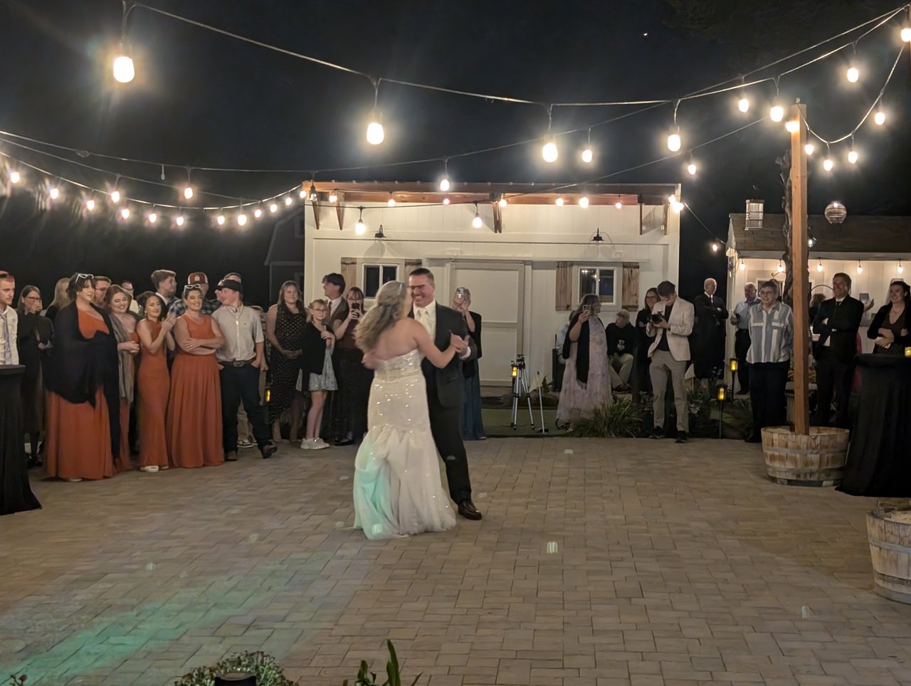 Emily and Logan slow dance under the string lights while the bridesmaids in rust gowns watch