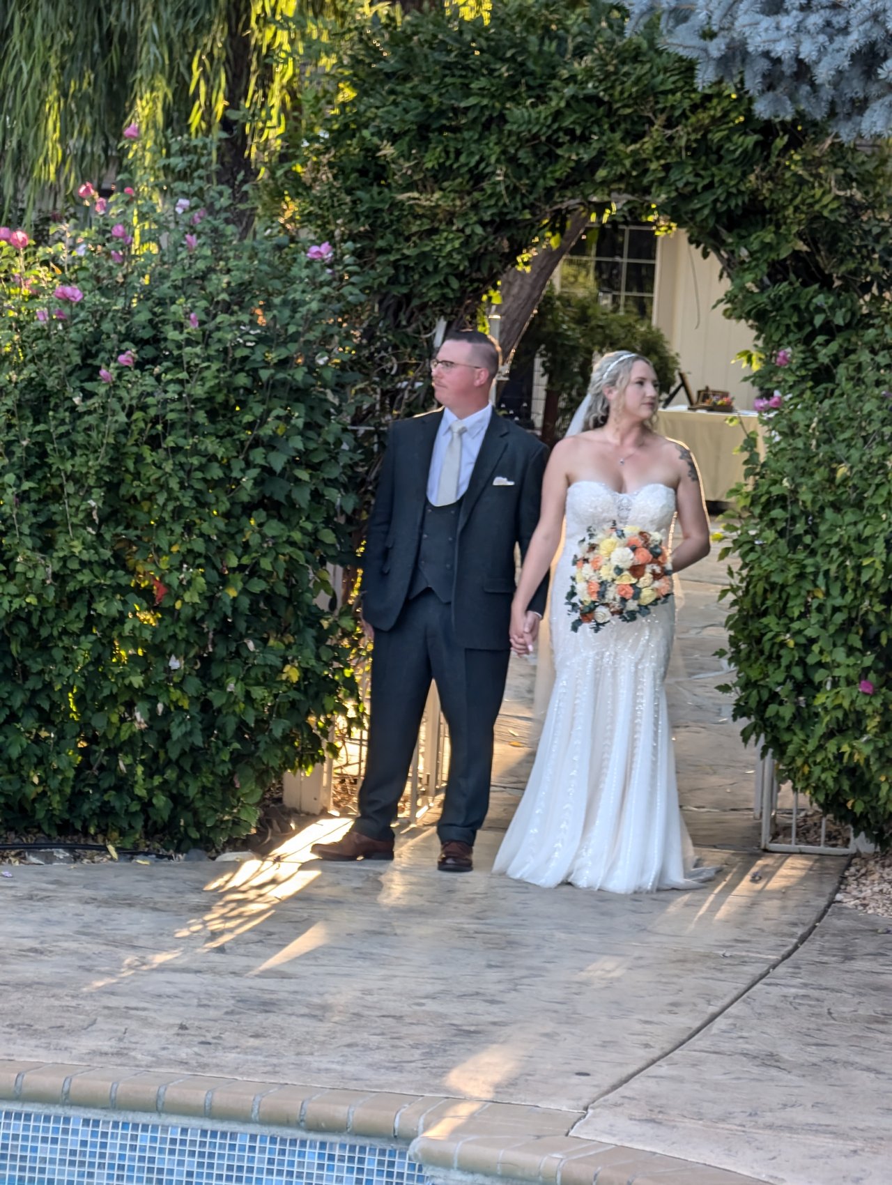 Emily and Logan stand together under the ivy arch at golden hour, her bouquet in hand