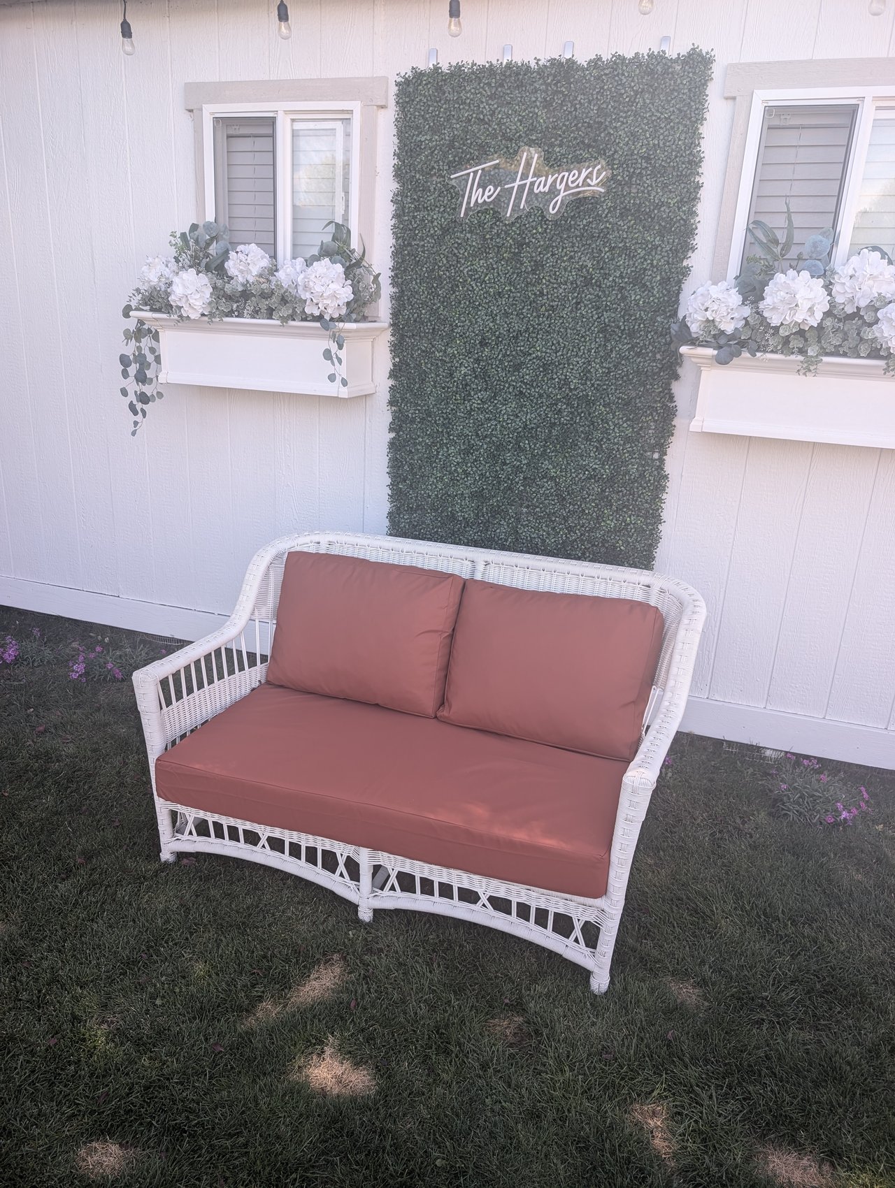 A neon sign reading The Hargers on a greenery wall above a white wicker loveseat with rust cushions