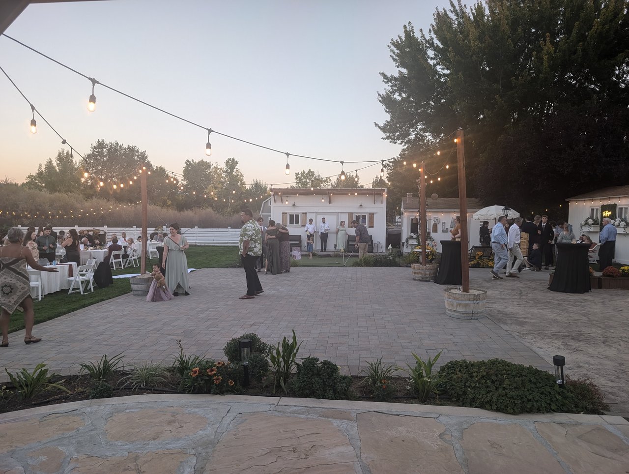 String lights glow over the patio dance floor at dusk as guests gather beside the white cottage