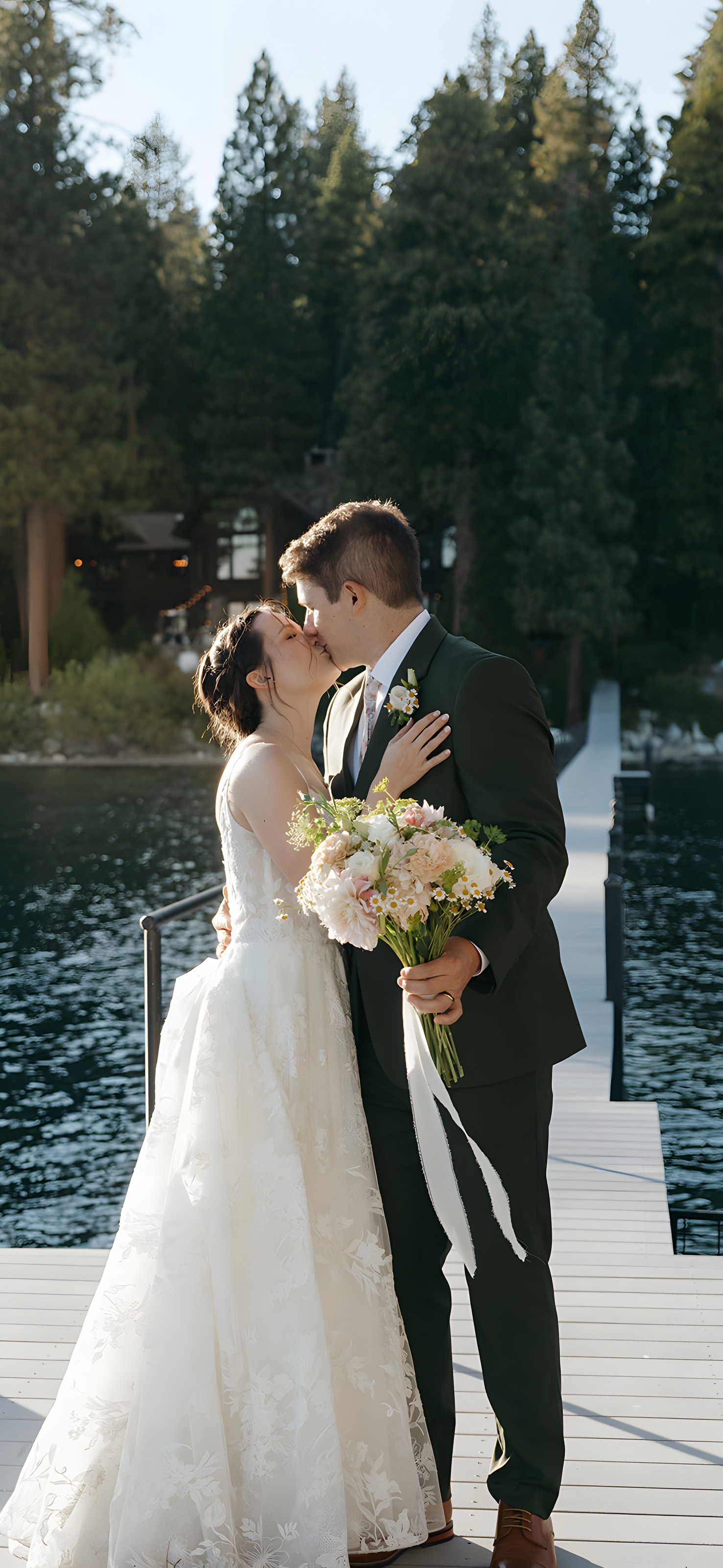 Emma and Jackson kiss on the dock at Lake Tahoe in Tahoe City, the pines and lakeside houses across the water behind them