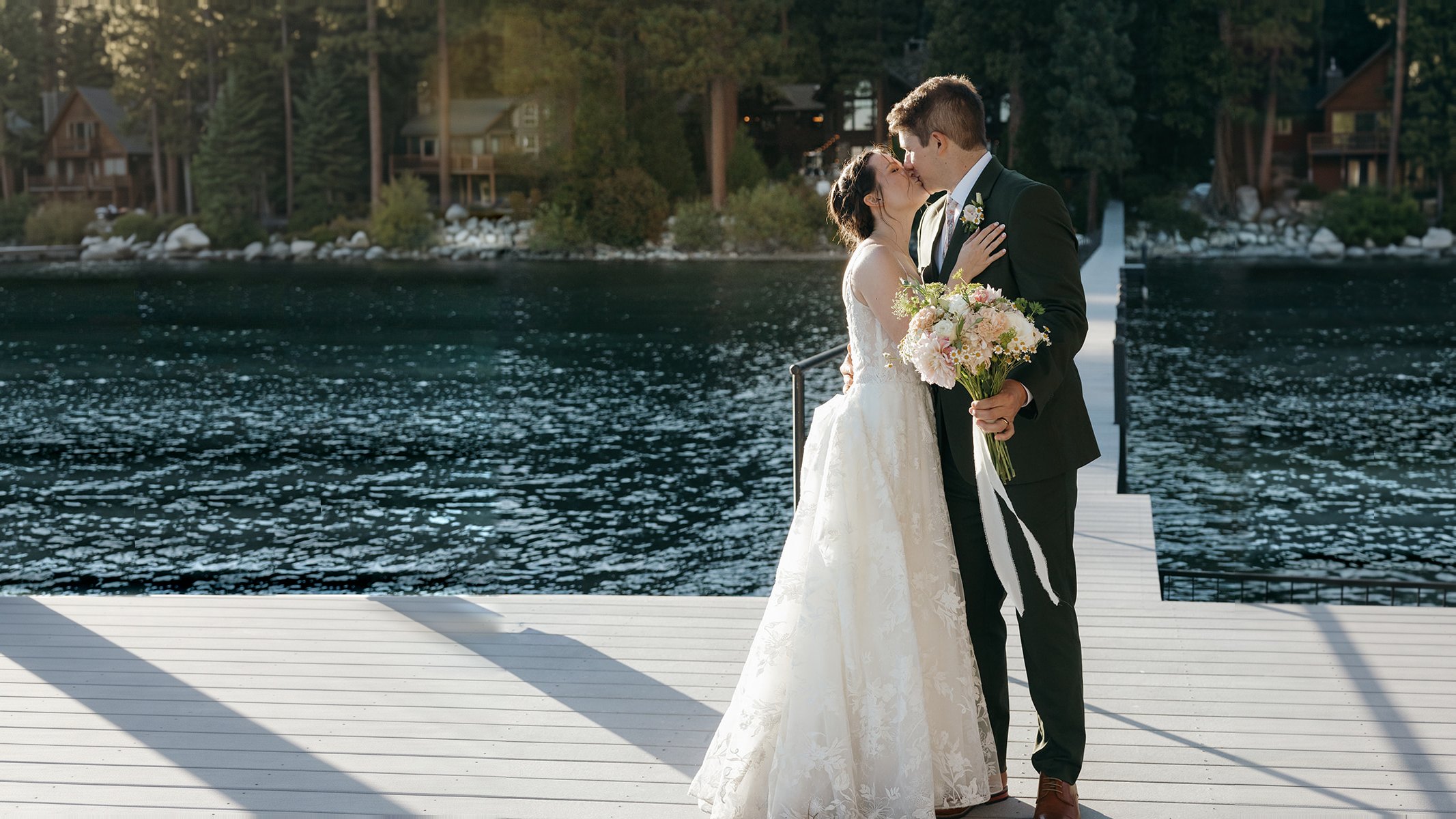 Emma and Jackson kiss on the dock at Lake Tahoe, her bouquet's white ribbons trailing, the pines and the house behind them