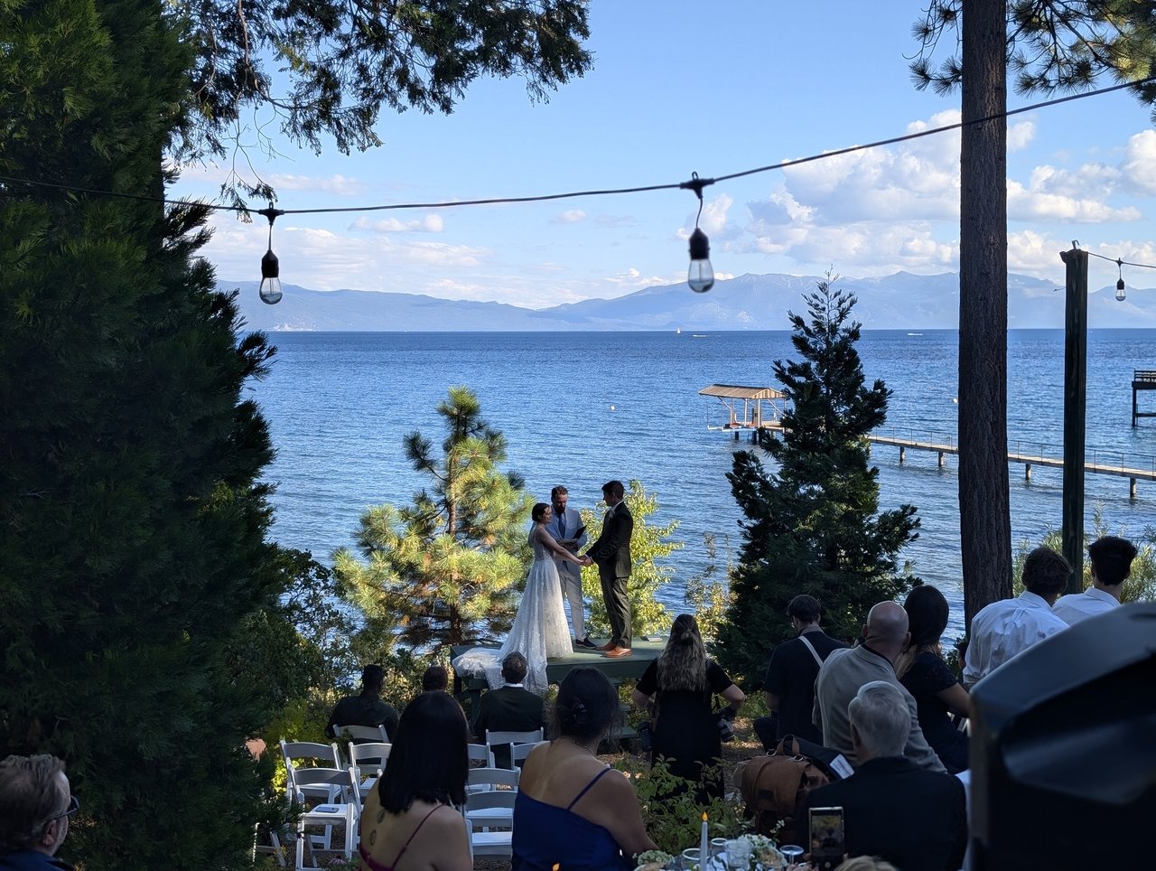 Emma and Jackson stand before the officiant as guests look on beneath the pines and string lights