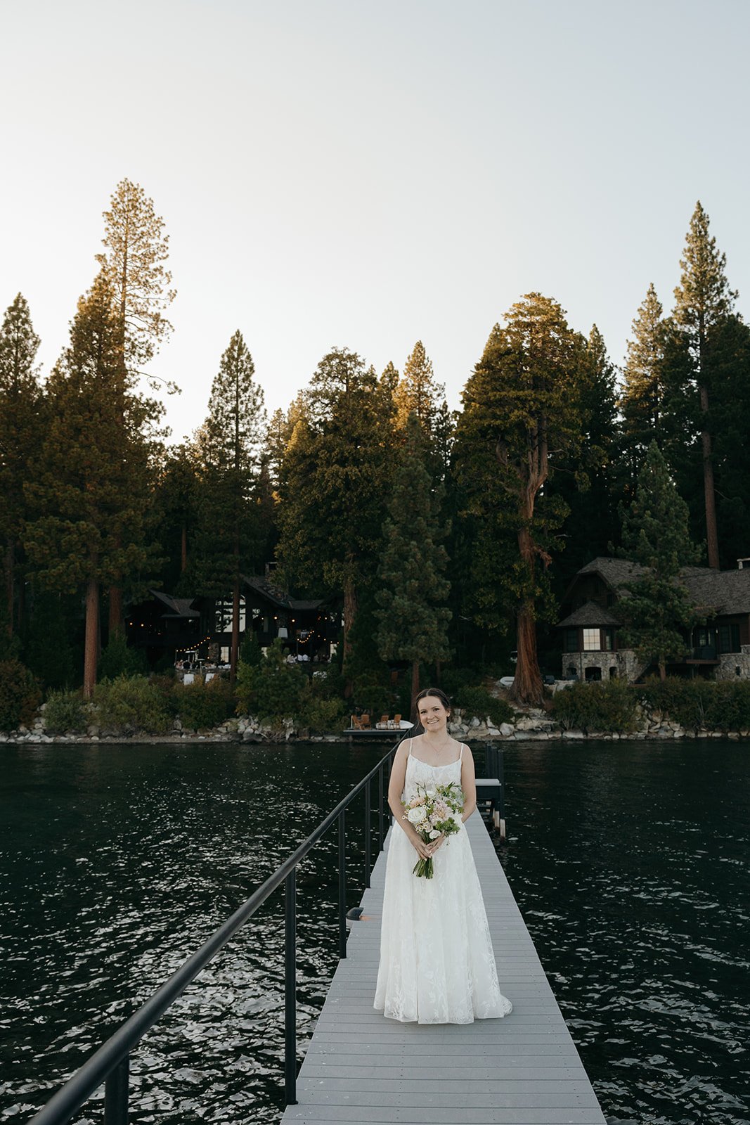 Emma stands on the long pier with her bouquet, the pines and lake houses behind her