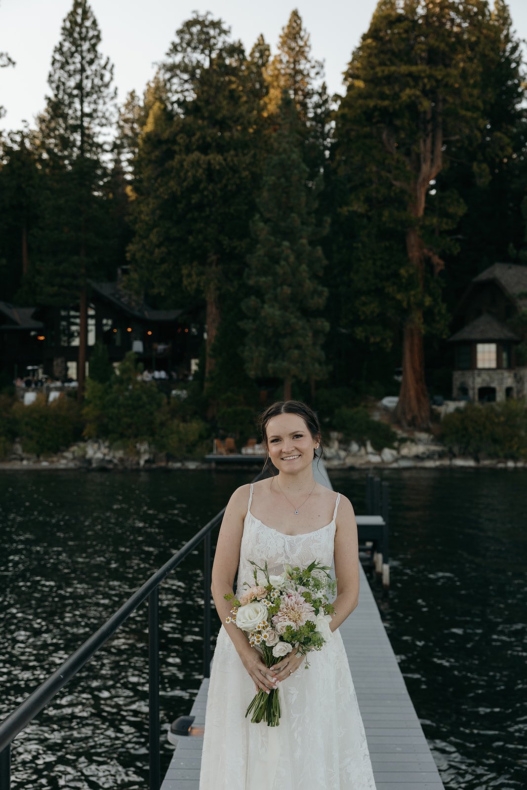 Emma smiles on the pier holding her bouquet, the pines behind her