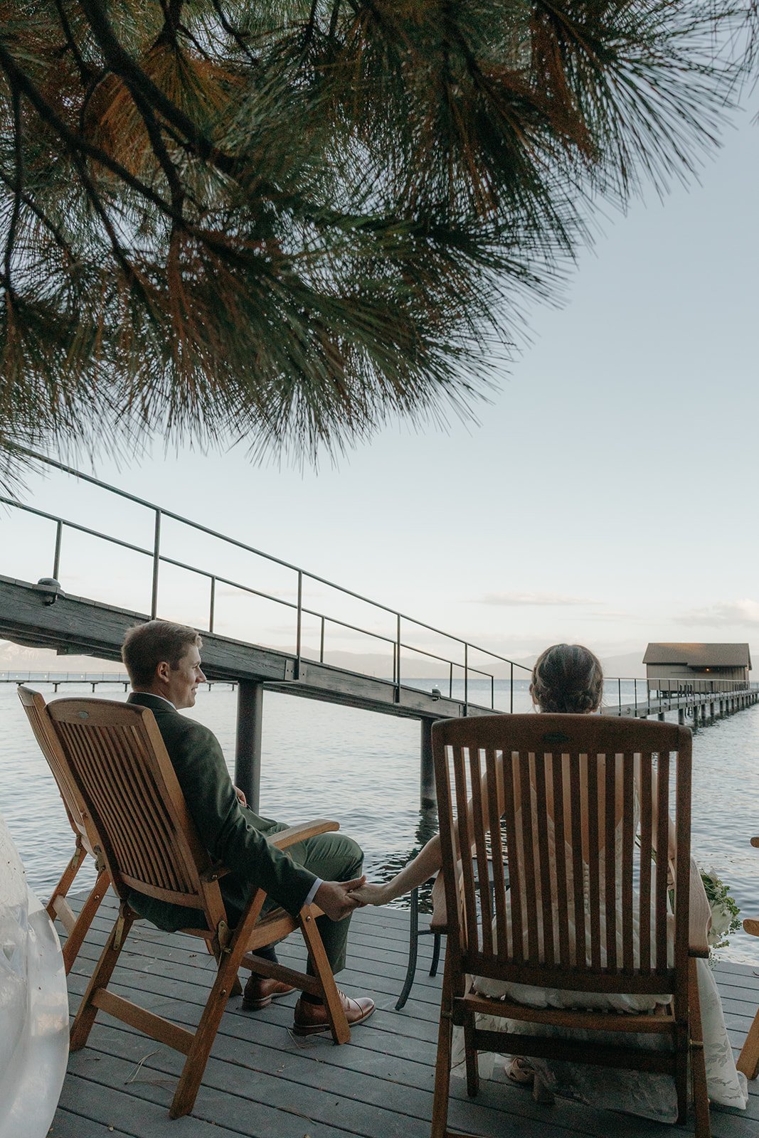 Emma and Jackson hold hands from two wooden chairs looking out over the lake