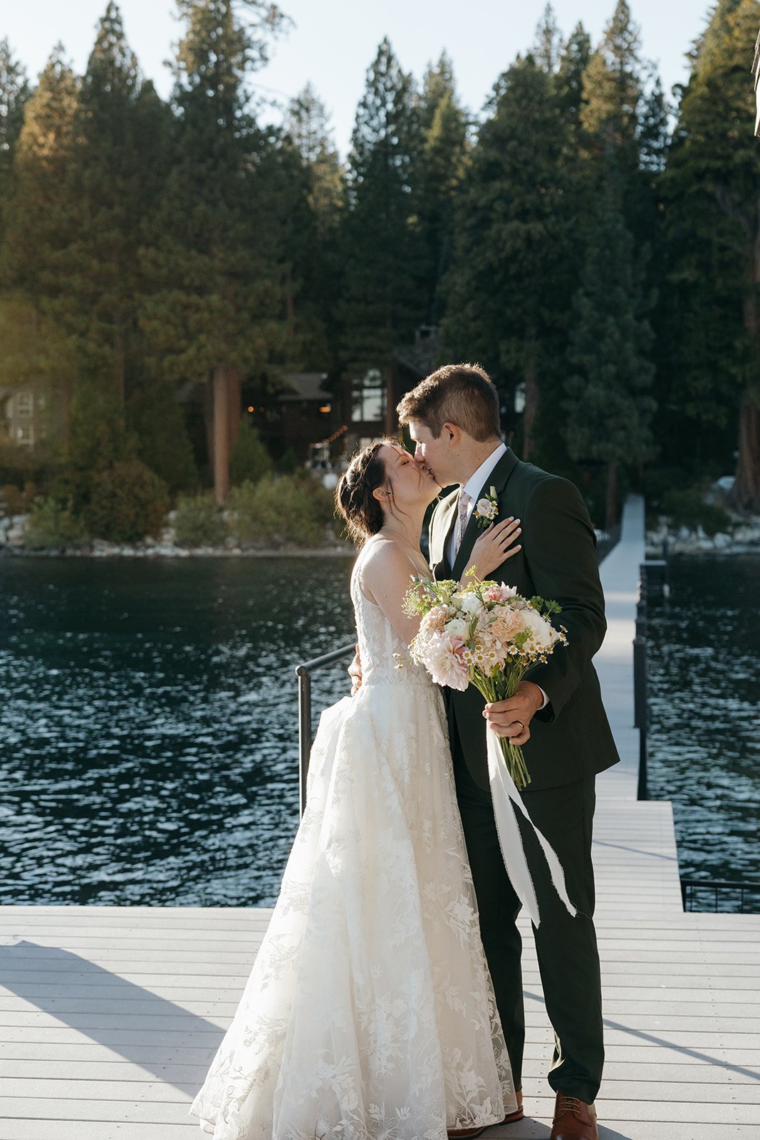 Emma and Jackson kiss on the dock at Lake Tahoe, her bouquet's white ribbons trailing, the pines and the house behind them