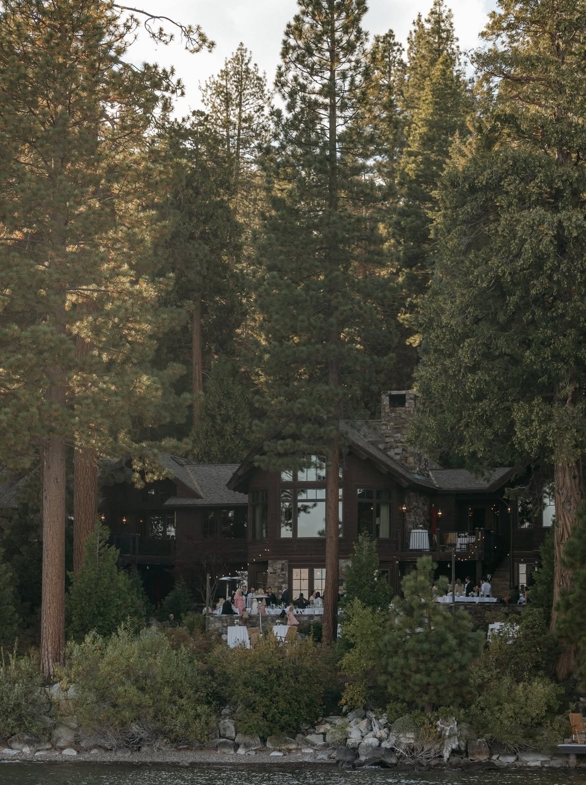 The house and the terrace party seen from across the water through the tall pines