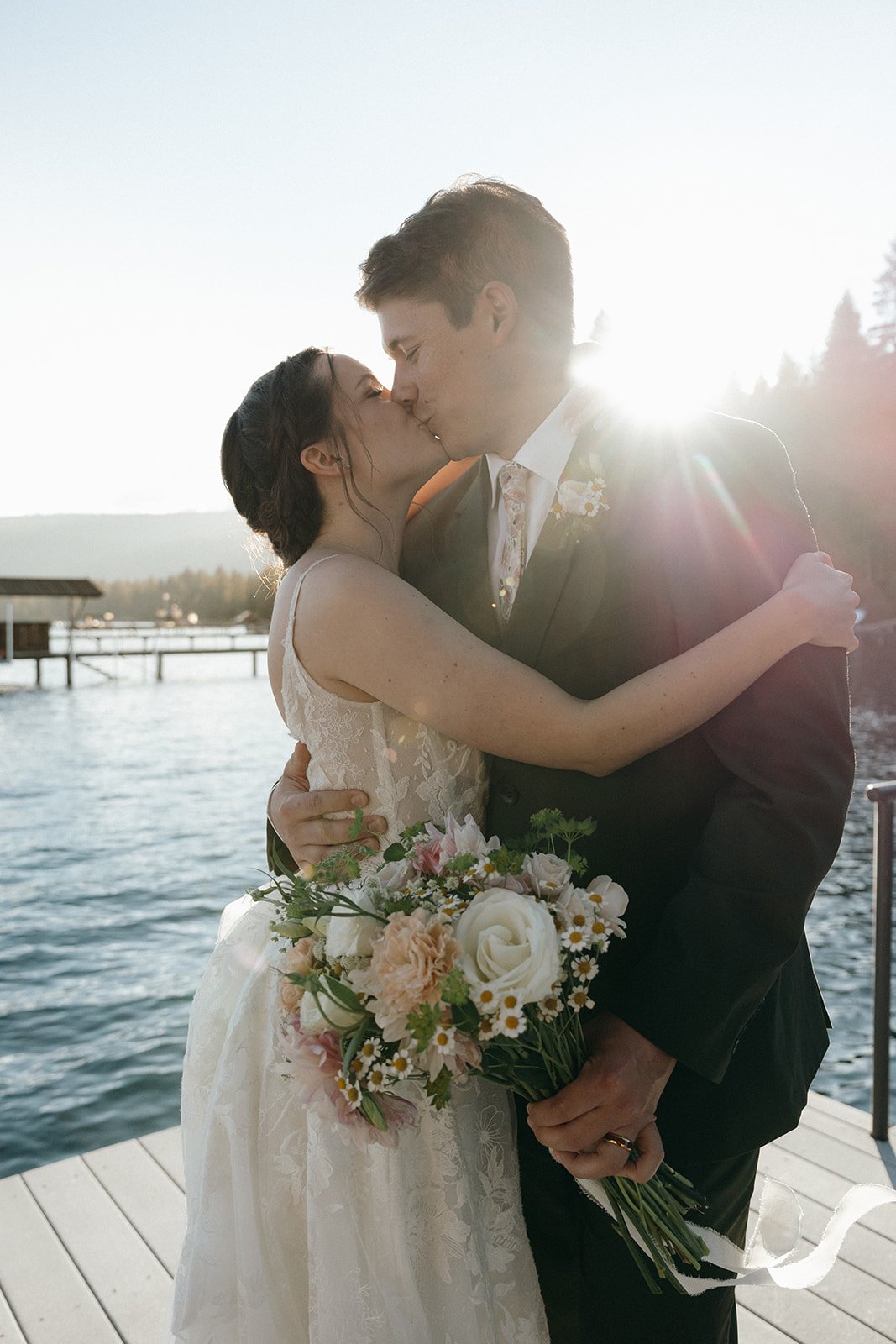 Emma and Jackson kiss in a warm embrace as the sun flares over the water