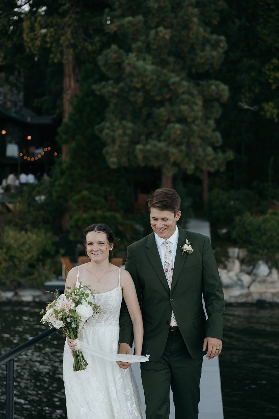 Emma and Jackson smile as they walk up from the dock, string lights glowing in the pines behind them