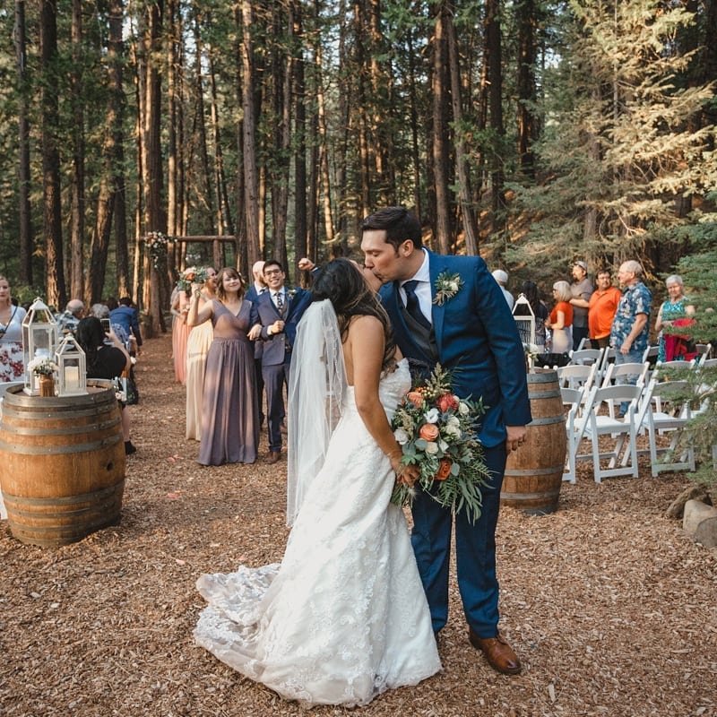 Hannah and Kyle kiss in the forest aisle between wine barrels and lanterns, photo by Composing Reality Photography
