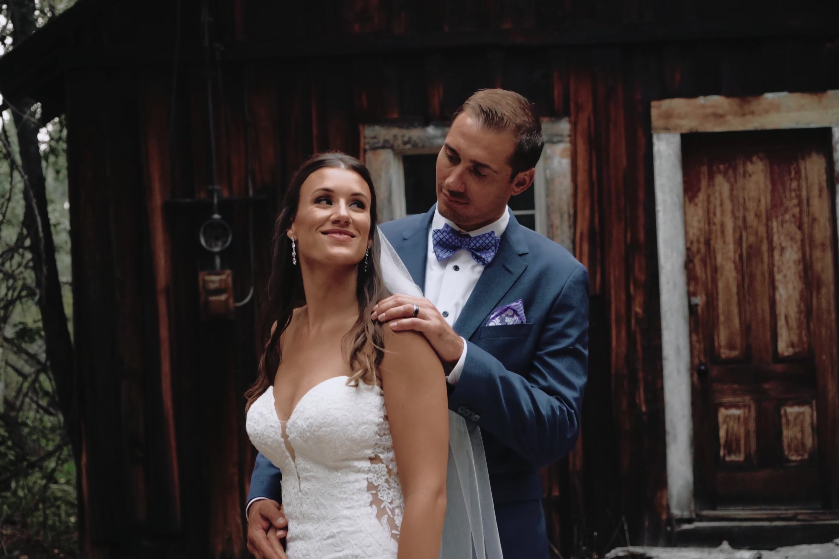 Wes rests his hands on Jen's shoulders in front of the old wooden cabin, a still from The Goodfolk Film Co. film