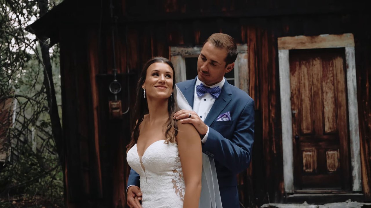 Wes rests his hands on Jen's shoulders in front of the old wooden cabin