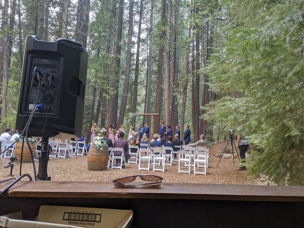 The forest ceremony from my DJ booth, guests in white chairs under the pines and the wedding party at the wooden arch