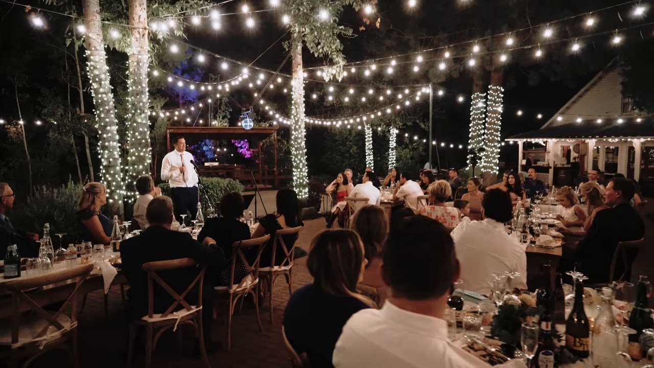 Guests listen to a toast at long tables under the string lights