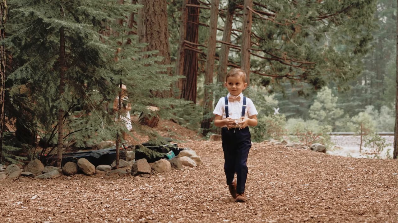 The ring bearer walks down the forest aisle in his suspenders