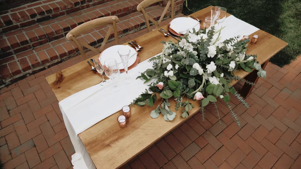 The sweetheart table with greenery and a white runner on the brick patio