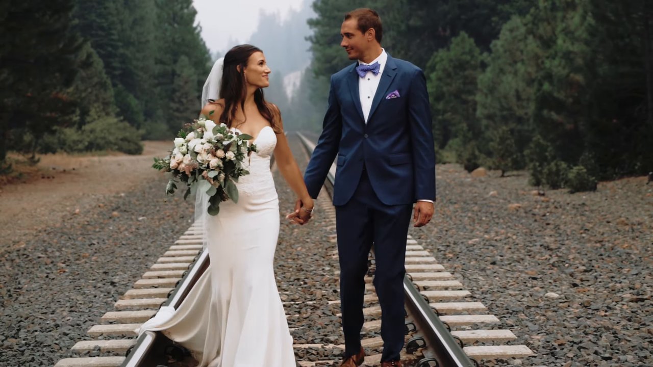 Jen and Wes walk hand in hand along the railroad tracks by the pines