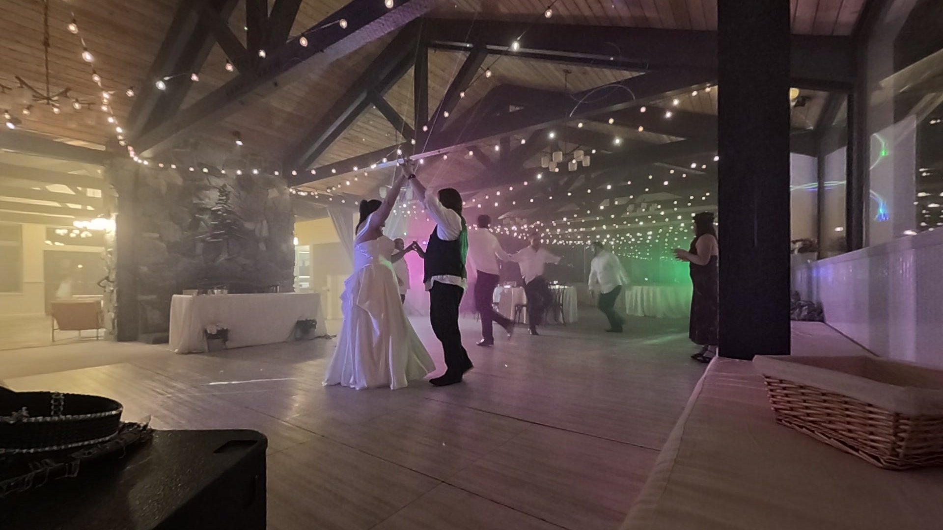 George raises Jericha's hand for a twirl on the dance floor at Tannenbaum Event Center, string lights overhead and pink and green light in the haze