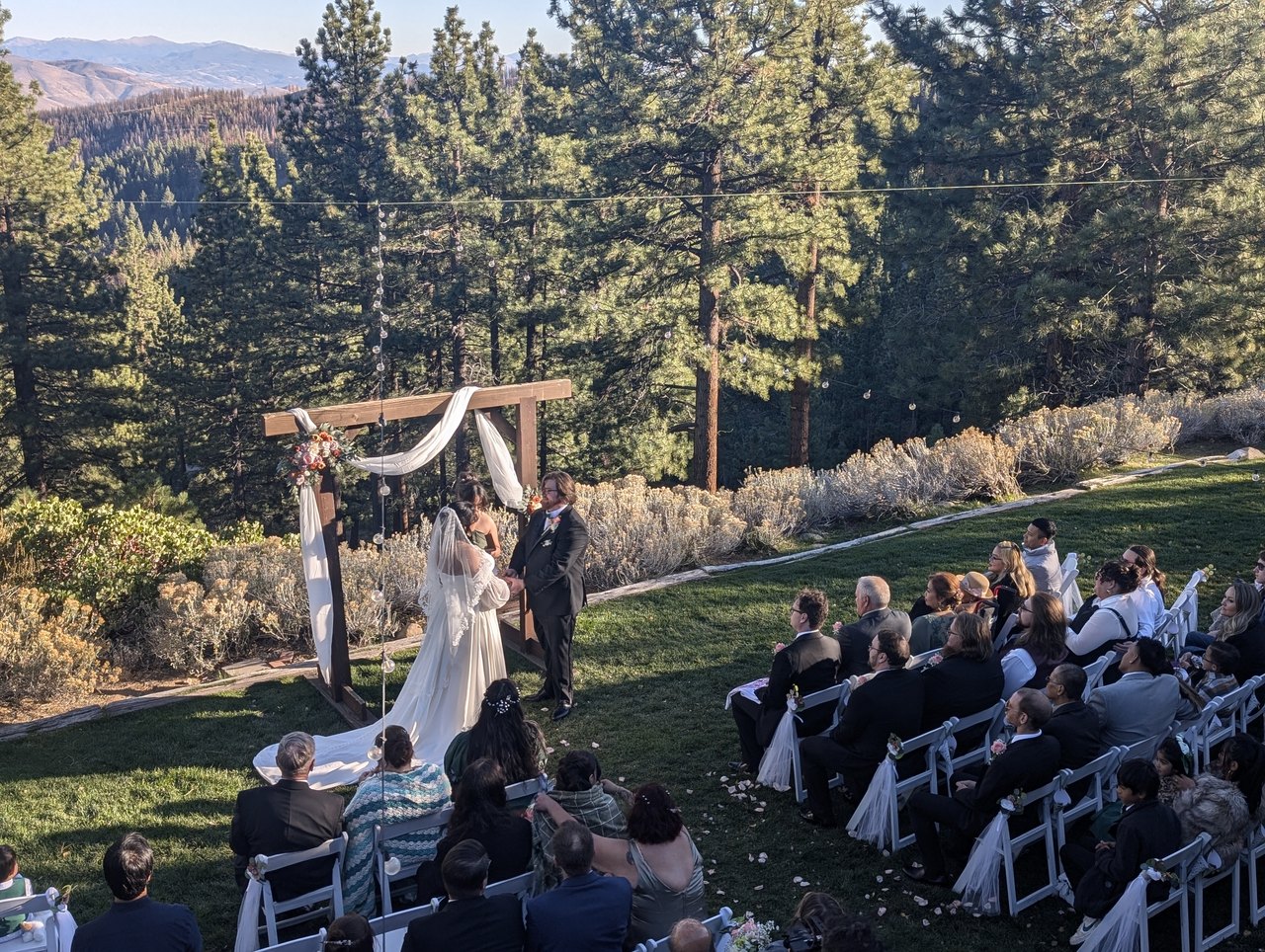 Jericha and George face each other at the arch under the tall pines at Tannenbaum as their guests look on