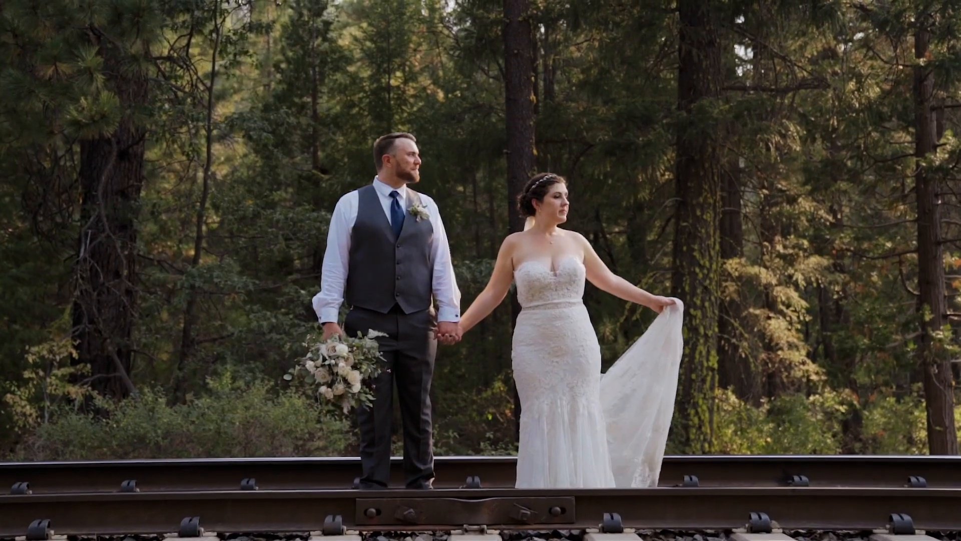 Jessie and Jeff stand hand in hand on the railroad tracks in the pines
