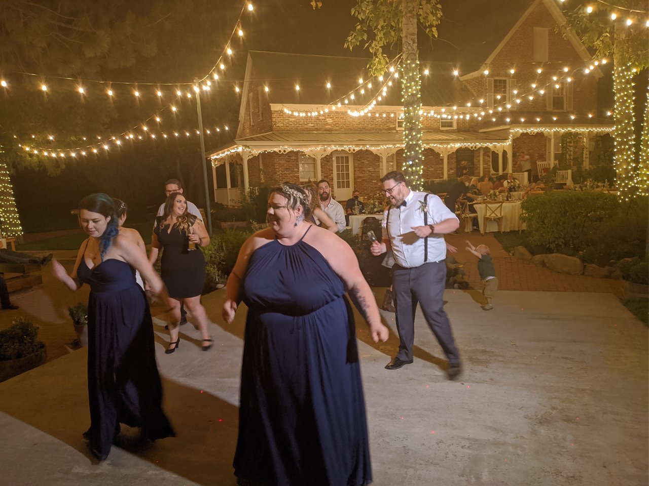 Bridesmaids in navy dresses dancing in front of the string lights and the brick house at Twenty Mile House
