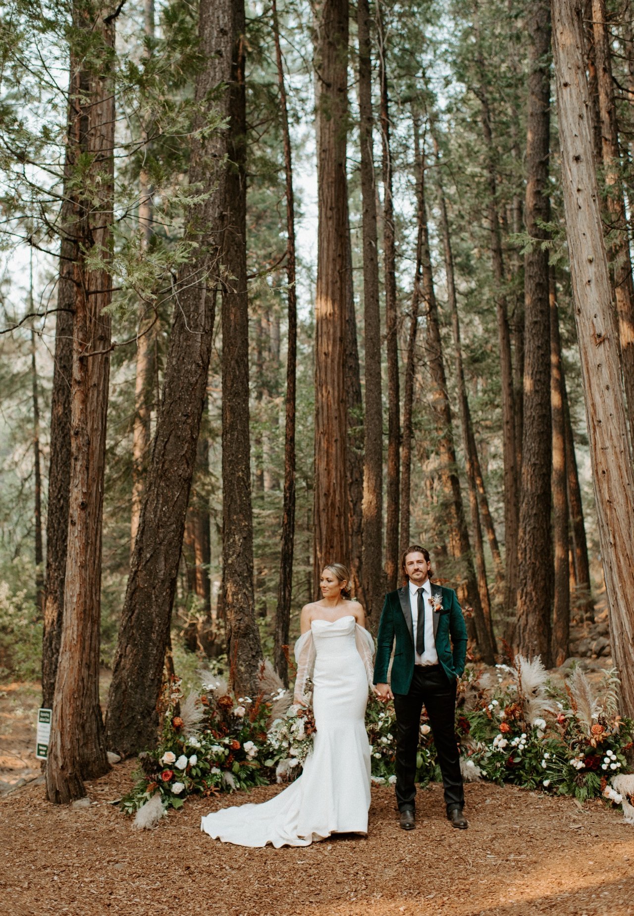 Joan and Kyle stand hand in hand at the altar among the tall trees