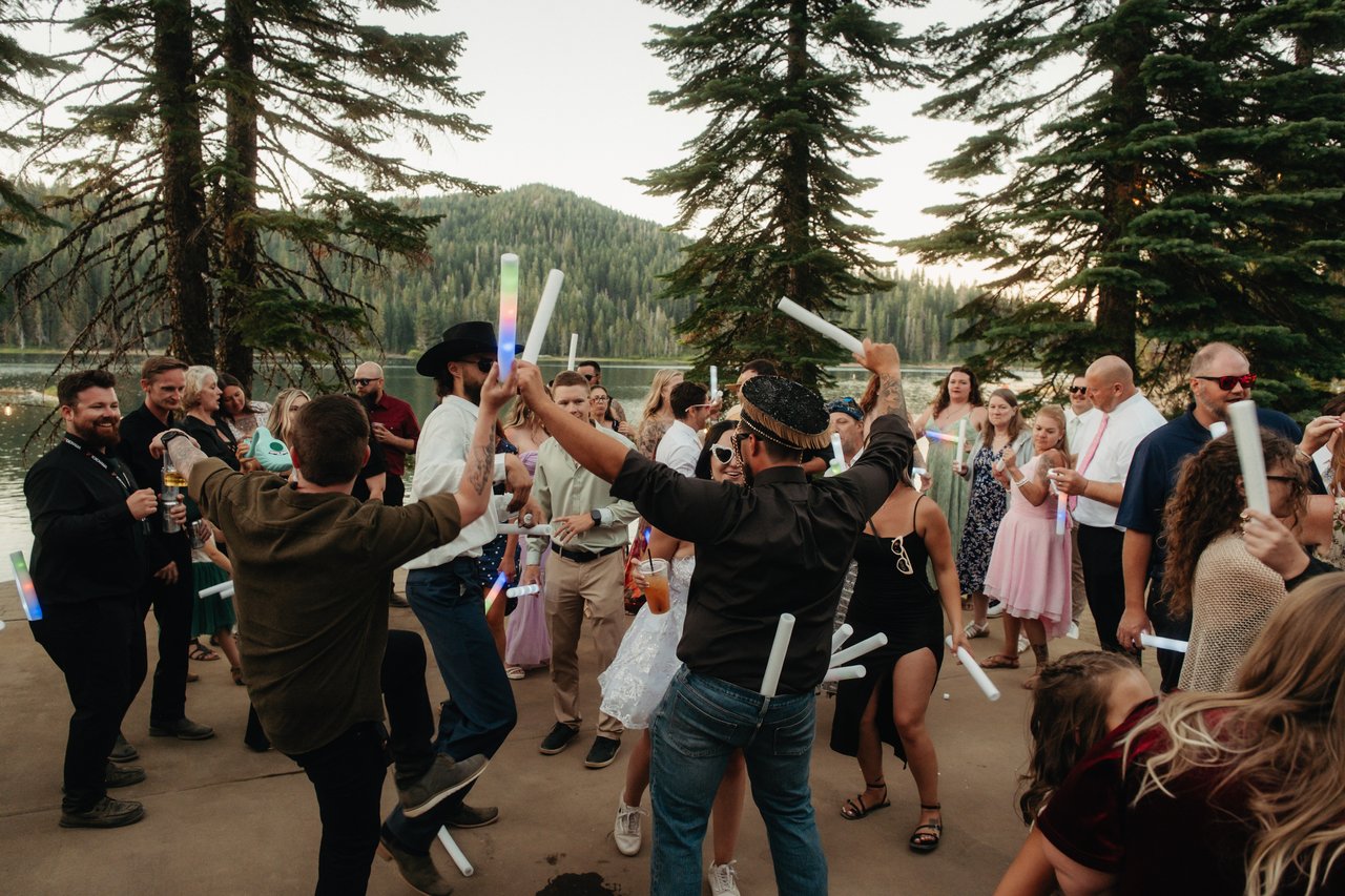 Guests dance in a circle beside Bucks Lake, raising glowing foam batons under the tall pines
