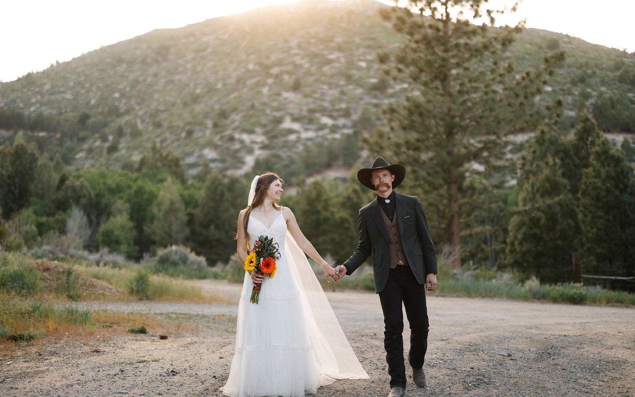 Kerstyn and Jakob walk hand in hand down the ranch road at golden hour, her sunflowers in hand