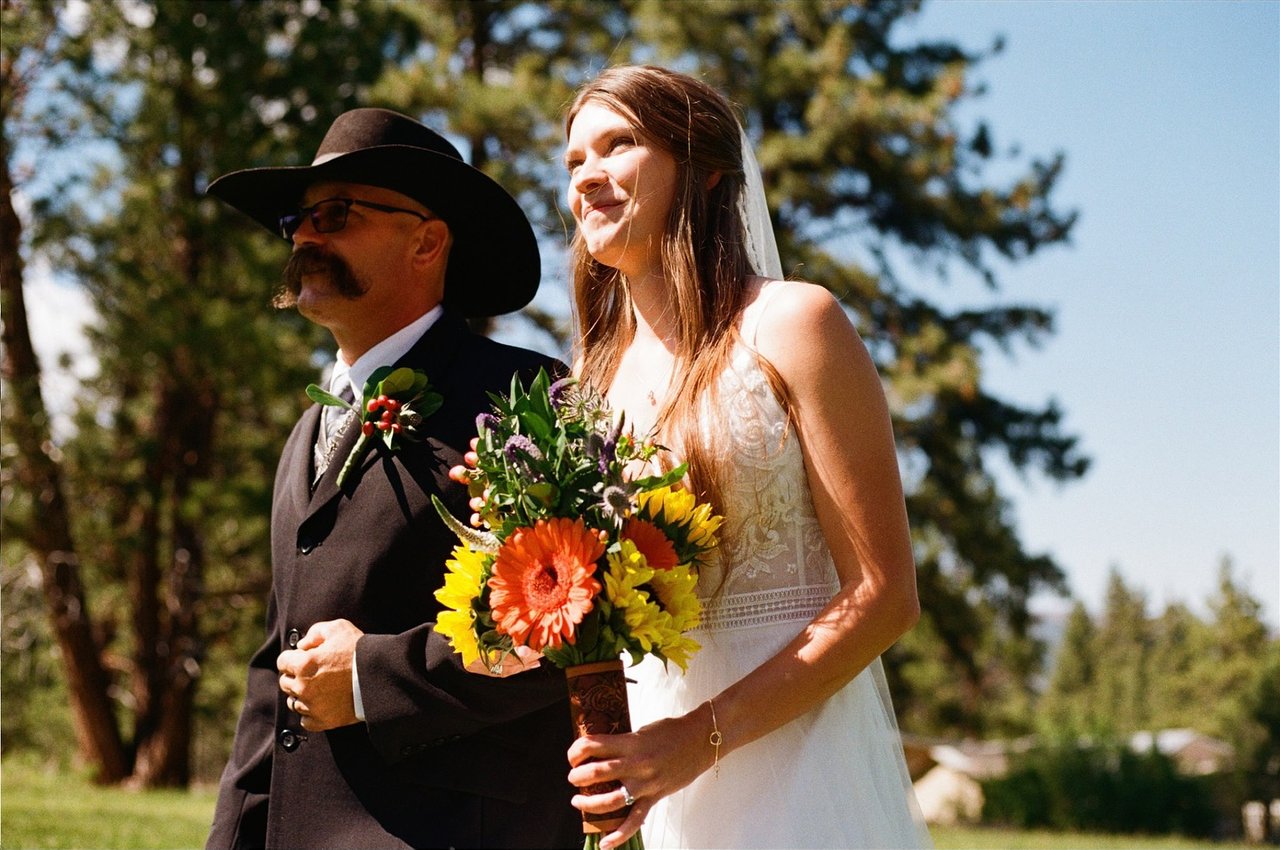 Kerstyn on her dad's arm under the pines, holding her sunflower bouquet